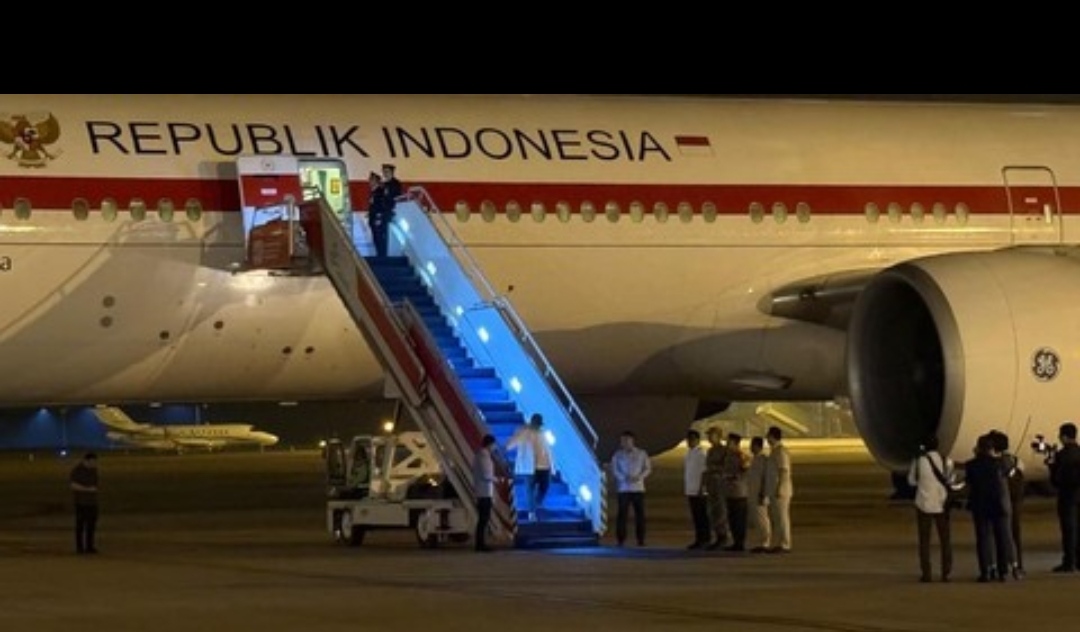 Pesawat kepresidenan bertuliskan Republik Indonesia parkir di landasan pacu pada malam hari saat seseorang menuruni tangga pesawat yang diterangi lampu biru. Di bawah tangga, sekelompok pejabat dan petugas berdiri berbaris menyambut kedatangan tersebut.