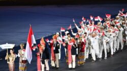 Barisan kontingen Indonesia melakukan defile di stadion, dipimpin pembawa papan nama dan bendera Merah Putih, diikuti rombongan atlet berseragam putih yang melambaikan bendera kecil.