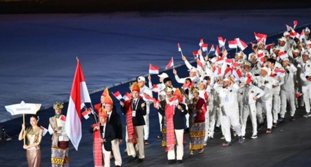 Barisan kontingen Indonesia melakukan defile di stadion, dipimpin pembawa papan nama dan bendera Merah Putih, diikuti rombongan atlet berseragam putih yang melambaikan bendera kecil.