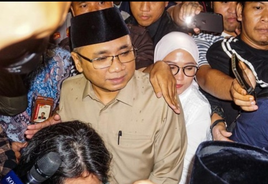 ​Mantan Menteri Agama Yaqut Cholil Qoumas mengenakan peci hitam dan kemeja cokelat berdesakan di tengah kerumunan awak media didampingi seorang wanita berkerudung putih.