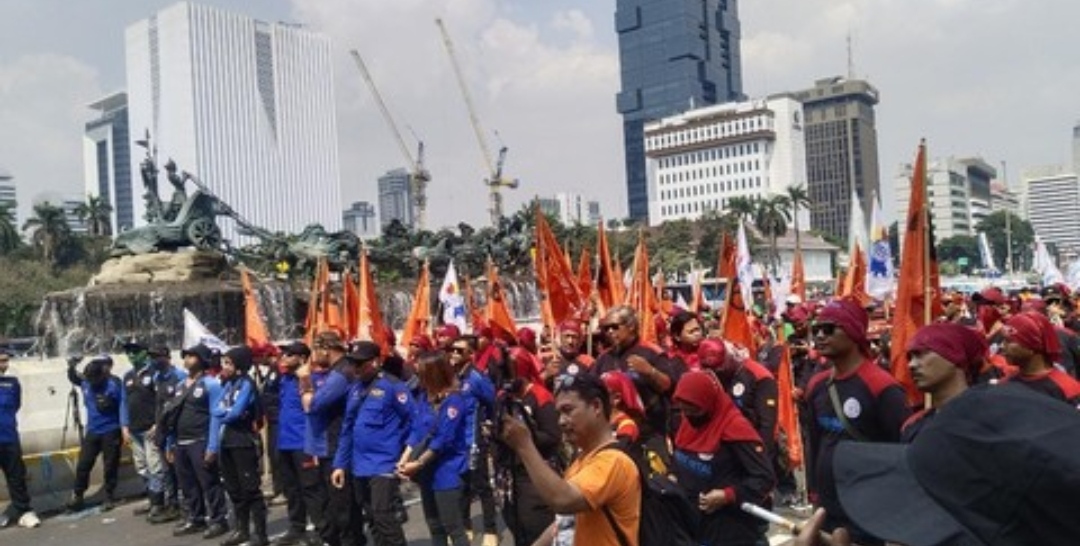 Sekelompok besar massa buruh berseragam merah-hitam membawa bendera oranye berkumpul di sekitar air mancur Patung Kuda Arjuna Wijaya, Jakarta, dengan latar belakang gedung bertingkat.