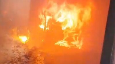 Petugas pemadam kebakaran menyiagakan armada darurat di jalan utama dekat sebuah gedung hijau sementara asap hitam pekat membubung tinggi dari atap rumah warga di area pemukiman padat.