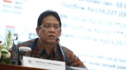 Menteri Keuangan Purbaya Yudhi Sadewa sedang berbicara dalam sebuah acara dengan latar belakang presentasi data keuangan. Ia mengenakan kemeja batik berwarna cokelat dan duduk di belakang meja yang bertuliskan namanya.