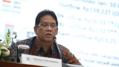 Menteri Keuangan Purbaya Yudhi Sadewa sedang berbicara dalam sebuah acara dengan latar belakang presentasi data keuangan. Ia mengenakan kemeja batik berwarna cokelat dan duduk di belakang meja yang bertuliskan namanya.