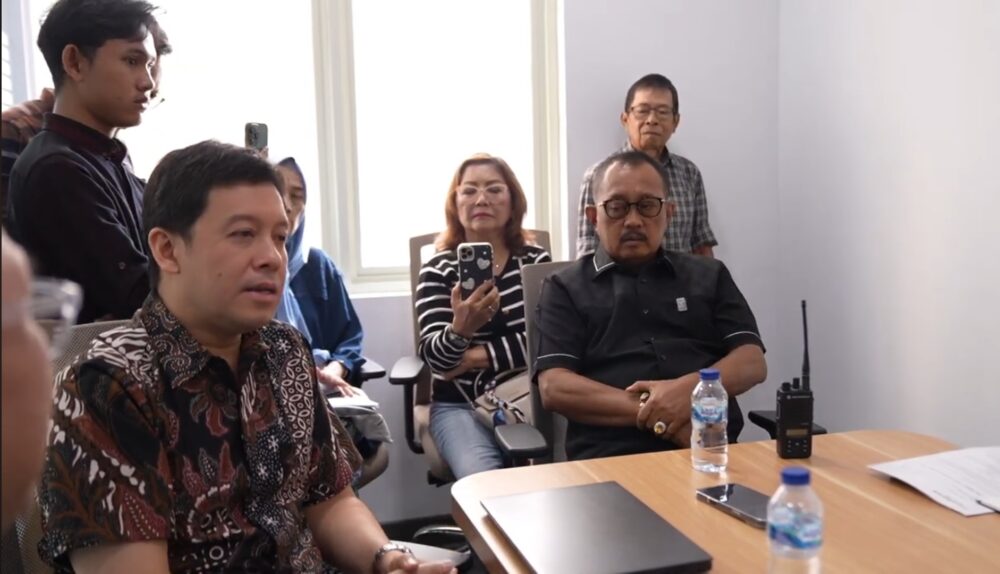 Foto menunjukkan Wakil Walikota Surabaya, Armuji, mengenakan kemeja hitam sedang memimpin mediasi di sebuah ruangan bersama beberapa orang lainnya. Di sisi kiri tampak seorang pria berbaju batik yang sedang berbicara, sementara di latar belakang terlihat beberapa warga yang memperhatikan jalannya diskusi di meja rapat yang berisi botol air mineral dan peralatan komunikasi.