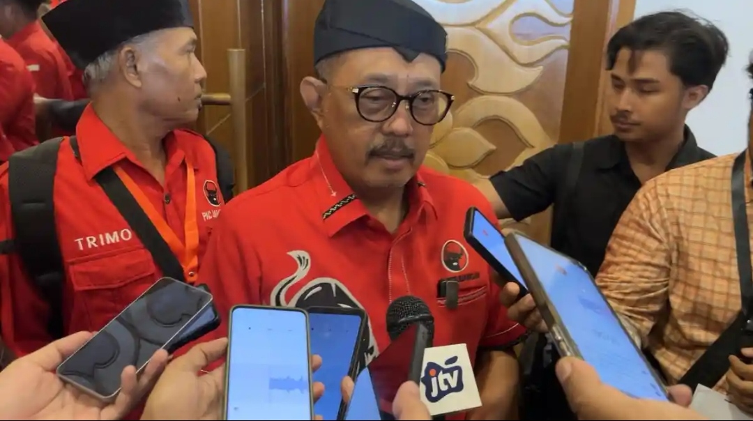 Seorang pria berkacamata mengenakan kemeja merah berlambang PDI Perjuangan dan udeng hitam sedang dikelilingi oleh awak media yang menyodorkan telepon genggam serta mikrofon untuk wawancara. Ekspresi wajahnya tampak serius saat memberikan keterangan di tengah kerumunan orang yang juga mengenakan seragam partai.
