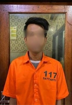 Seorang pria tersangka pencurian mengenakan baju tahanan oranye dengan nomor 117 dan tulisan "SATTAHTI" di bagian dada, berdiri di depan pintu kayu dengan latar belakang dinding bermotif.