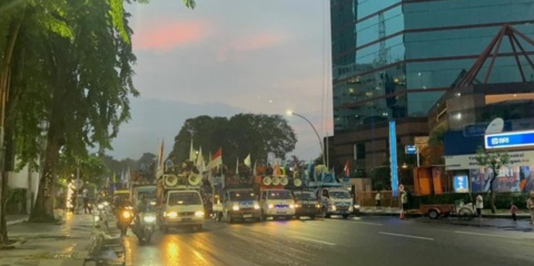 Deretan truk berkonvoi membawa massa aksi buruh melintas di jalan utama Kota Surabaya saat senja, dilengkapi pengeras suara dan bendera, dengan lalu lintas tampak tersendat di sekitarnya.