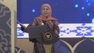 Seorang pejabat perempuan yang menjabat sebagai gubernur Jawa Timur berdiri di belakang podium berlogo negara, menyampaikan sambutan dalam sebuah acara resmi dengan latar layar besar bernuansa biru.