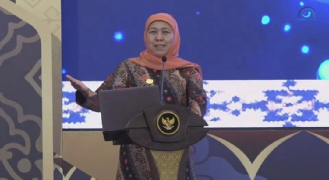 Seorang pejabat perempuan yang menjabat sebagai gubernur Jawa Timur berdiri di belakang podium berlogo negara, menyampaikan sambutan dalam sebuah acara resmi dengan latar layar besar bernuansa biru.