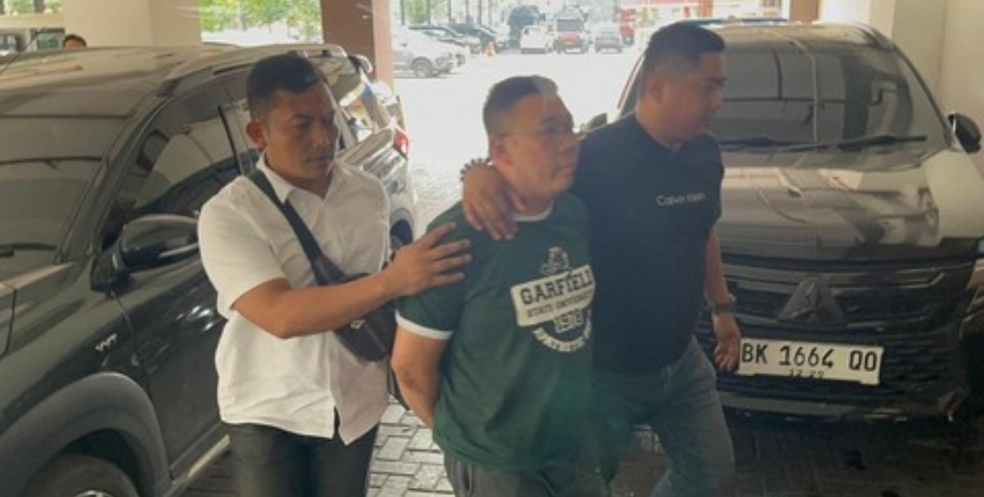 Seorang pria berbaju hijau digiring dua petugas kepolisian dengan tangan terborgol di area parkir gedung, dikelilingi kendaraan yang terparkir.