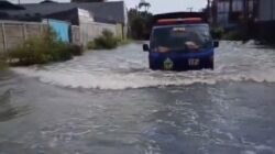 Sebuah truk operasional milik BPBD Surabaya dengan logo 112 melintasi jalan yang tergenang banjir cukup tinggi di kawasan permukiman yang dibatasi tembok pagar.
