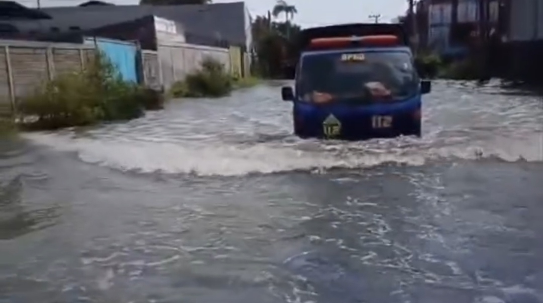 Sebuah truk operasional milik BPBD Surabaya dengan logo 112 melintasi jalan yang tergenang banjir cukup tinggi di kawasan permukiman yang dibatasi tembok pagar.