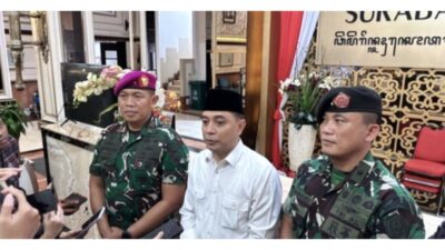 Seorang pejabat pemerintah berdiri di tengah, didampingi dua personel TNI berseragam, sedang memberikan keterangan kepada wartawan di dalam gedung Balai Kota Surabaya.