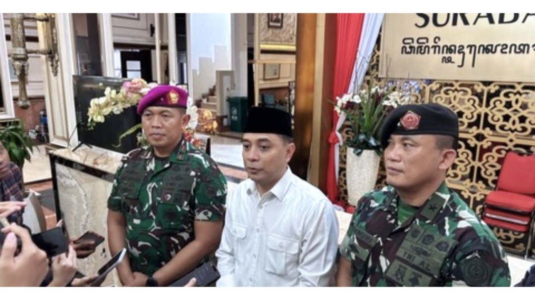 Seorang pejabat pemerintah berdiri di tengah, didampingi dua personel TNI berseragam, sedang memberikan keterangan kepada wartawan di dalam gedung Balai Kota Surabaya.