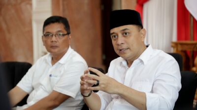 Wali Kota Surabaya Eri Cahyadi yang mengenakan kemeja putih dan peci hitam tampak sedang berbicara sambil memberikan gestur tangan, didampingi seorang pria berkacamata di sebuah ruangan berlatar bendera Merah Putih.