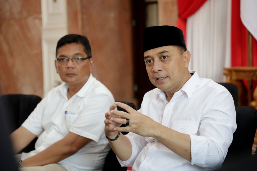 Wali Kota Surabaya Eri Cahyadi yang mengenakan kemeja putih dan peci hitam tampak sedang berbicara sambil memberikan gestur tangan, didampingi seorang pria berkacamata di sebuah ruangan berlatar bendera Merah Putih.