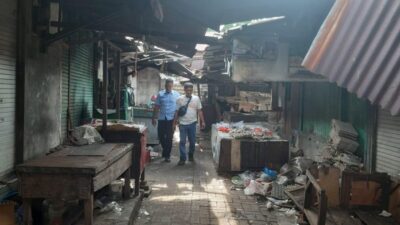 Dua orang pria berjalan melewati lorong pasar tradisional yang tampak kumuh dengan bangunan kayu yang mulai rusak.