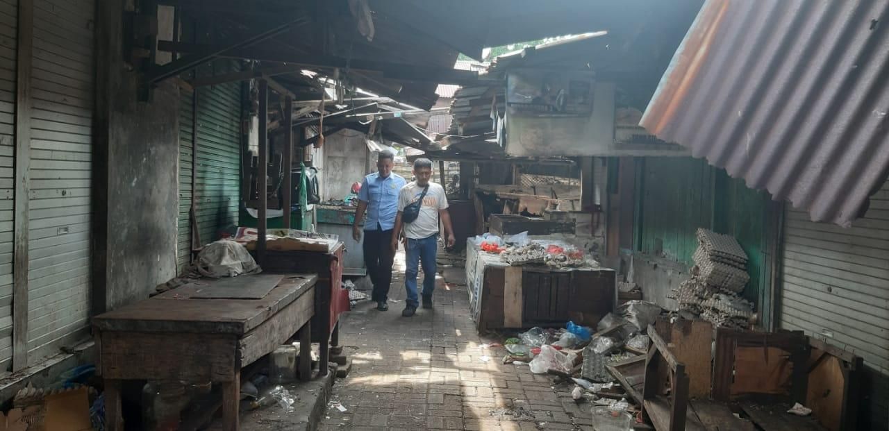 Dua orang pria berjalan melewati lorong pasar tradisional yang tampak kumuh dengan bangunan kayu yang mulai rusak.
