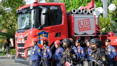 Barisan petugas pemadam kebakaran berseragam biru dengan helm merah dan petugas Satpol PP berseragam hitam dengan sepeda motor bersiaga di depan truk pemadam kebakaran Scania berwarna merah milik Pemerintah Kota Surabaya yang bertuliskan layanan darurat 112.