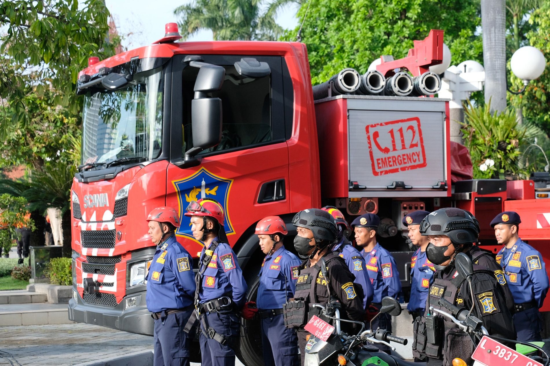 Barisan petugas pemadam kebakaran berseragam biru dengan helm merah dan petugas Satpol PP berseragam hitam dengan sepeda motor bersiaga di depan truk pemadam kebakaran Scania berwarna merah milik Pemerintah Kota Surabaya yang bertuliskan layanan darurat 112.