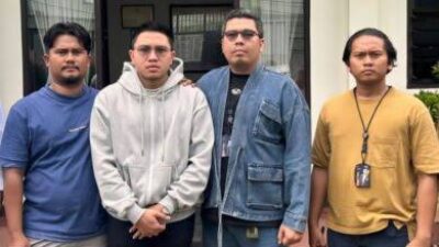 Empat pria berdiri berjejer di depan sebuah bangunan, dengan dua pria di tengah mengenakan hoodie dan jaket denim.