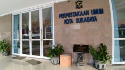 Tampak pintu masuk Perpustakaan Umum Kota Surabaya dengan papan nama terpasang di dinding, pintu kaca, serta pot tanaman di sisi kanan dan kiri area lobi.