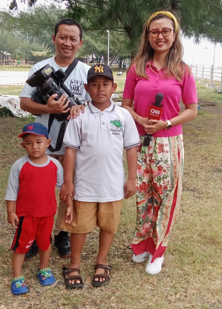 Seorang reporter wanita memegang mikrofon merah berlogo CNN Indonesia dan juru kamera pria berfoto bersama dua anak laki-laki di area taman terbuka dengan latar pepohonan.
