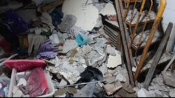 Puing beton, pecahan dinding, dan perabot rumah tangga terlihat berserakan di dalam ruangan akibat ambruknya struktur lantai, dengan besi tulangan dan rak kayu masih berdiri di sisi kanan gambar.