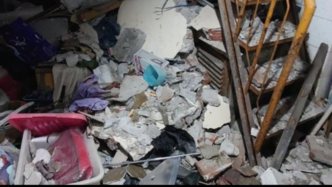 Pu­ing beton, pecahan dinding, dan perabot rumah tangga terlihat berserakan di dalam ruangan akibat ambruknya struktur lantai, dengan besi tulangan dan rak kayu masih berdiri di sisi kanan gambar.