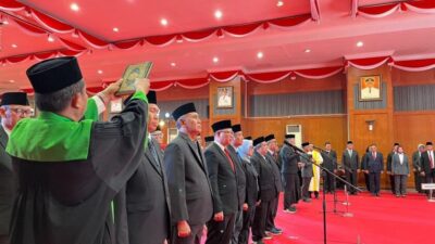 Seorang rohaniwan mengangkat kitab suci Al-Qur'an di atas kepala pejabat pria berjas hitam saat prosesi pengambilan sumpah jabatan di aula berkarpet merah.