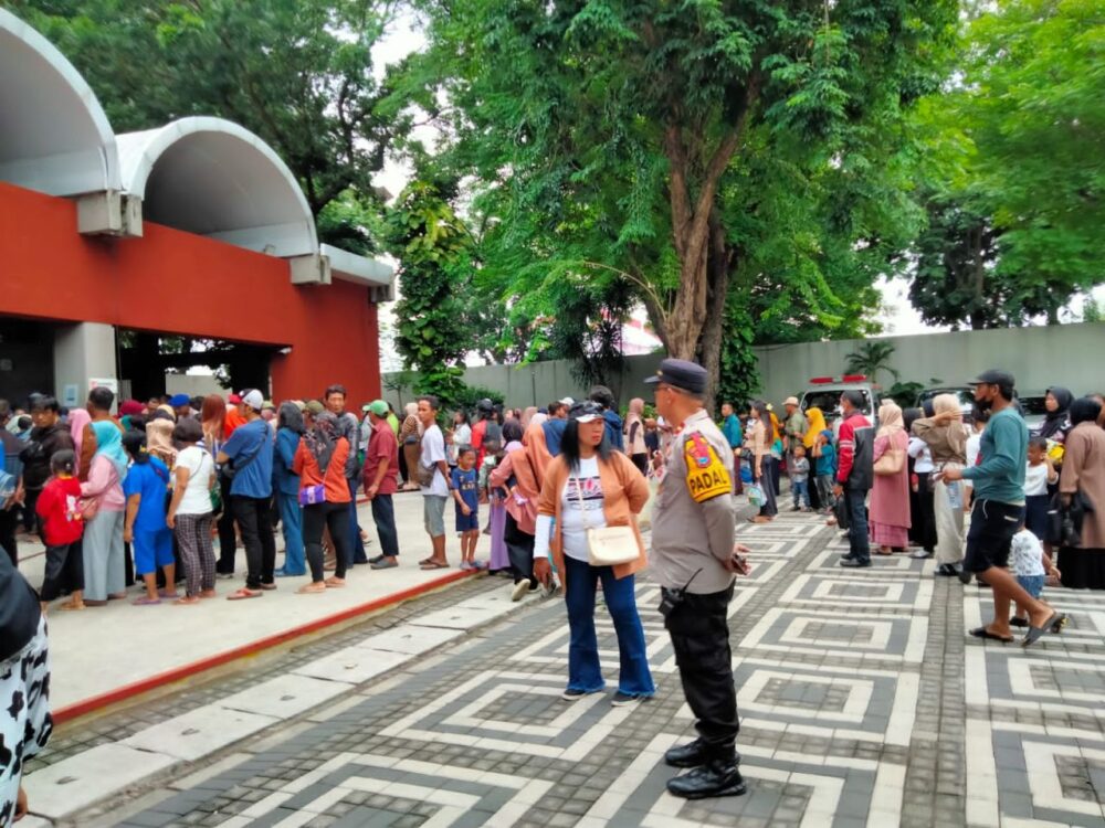 Kerumunan pengunjung berdiri mengantre di area berlantai paving depan bangunan merah, sementara seorang petugas polisi berseragam lengkap berdiri memantau situasi di sisi kanan.