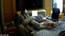 Seorang wanita duduk di tempat tidur di dalam kamar, sementara seorang pria berdiri di dekat lemari di latar belakang, seperti terlihat dalam tangkapan layar video dengan stempel waktu.